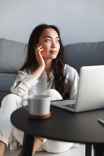 Vertical Shot Of Dreamy Attractive Asian Girl Sitting Near Laptop At Home And Looking Outside Window With Romantic Gaze. Woman Resting In Her Apartment With Cup Of Coffee And Computer