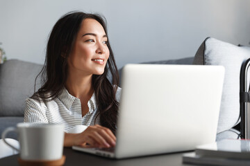 Close-up of ambitious beautiful asian female employee working from home. Girl on remote using laptop, smiling dreamy while looking outside window. Student feel inspired writing essay