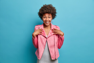 Canvas Print - Half length shot of pretty cheerful Afro American girl dressed in fashionable pink jacket, smiles broadly, hears exciting good news, poses against blue background. Emotions, feelings concept