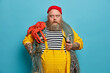 © wayhome.studio  - Stupefied bearded man stares bugged eyes at camera holds pipe dressed in yellow overalls red hat carries big sea creature on shoulder, poses against blue background, busy with maintenance of ship