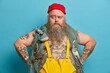 © wayhome.studio  - Photo of serious bearded man has confident expression keeps hands on hips, poses with fishing gears, dressed in overalls, red hat, goes agling, poses against blue background. Sea adventure concept