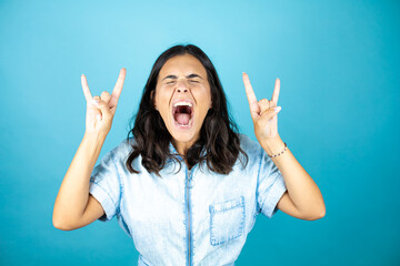 Young beautiful woman wearing a denim jumpsuit over isolated blue background shouting with crazy expression doing rock symbol with hands up.