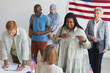 © Seventyfour - Multi-ethnic group of people registering at polling station decorated with American flags on election day, focus on smiling African woman pointing at I VOTE sticker, copy space