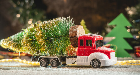 Wall Mural - A red and white toy truck is transporting a Christmas tree against a background of lights and paper pines and snow