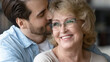© fizkes - Close up loving young man kissing smiling beautiful mature mother wearing glasses, hugging from back, standing at home, overjoyed happy middle aged woman and adult son enjoying tender moment