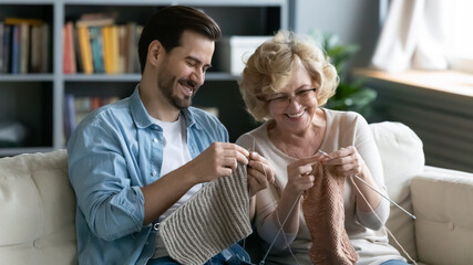 Wall Mural - Happy mature mother and son knitting together, enjoying leisure time at home, smiling middle aged woman wearing glasses and young man sitting on cozy couch in living room, holding needles