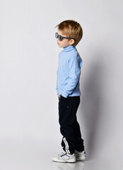 Wall Mural - Self-confident blond kid boy in sneakers, black sport pants, blue turtleneck sweater and sunglasses stands side to us