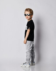 Wall Mural - Blond kid boy in black t-shirt, gray pants and sneakers stands side to us with head turned and looks at camera