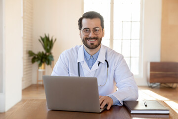 Wall Mural - Head shot portrait smiling doctor wearing glasses working on laptop, sitting at desk in office, therapist physician gp wearing white medical uniform with stethoscope looking at camera