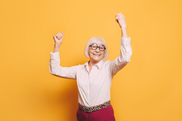 Happy elderly woman with white hair, round glasses wearing white blouse, red pants and leopard print belt standing isolated over orange background with raised arms.