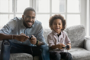 Happy young african ethnicity father sitting on sofa, involved in funny video game with laughing cute small kid son at home, emotional multiracial family enjoying competition with controllers in hands