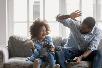 Wall Mural - Overjoyed african american family involved in video game competition at home, excited emotional young mixed race father giving high five to laughing adorable small child daughter, celebrating win.