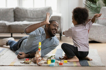 Happy young handsome biracial father lying on floor carpet, giving high five to joyful little preschool child son, finishing constructing building, cheerful multiracial family having fun on weekend.