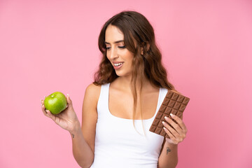 Wall Mural - Young woman over isolated pink background taking a chocolate tablet in one hand and an apple in the other