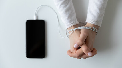 Top above close up view female hands lying on table wrapped in wires cables from electronic gadget smartphone, young generation addiction to social networks, modern technology obsession concept.