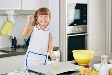 Wall Mural - Portrait of happy preteen girl covering baking sheet with soft butter before putting dough on it