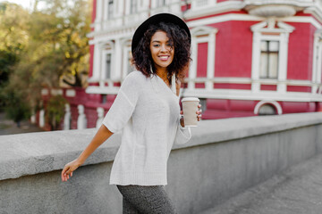 Wall Mural - Playful woman wraps herself over her shoulder and looking in camera. Wearing. Stylish hipster outfit. Afro hairstyle . Perfect spring  leisure time in the evening City.