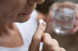 Close up senior mature woman taking painkiller, antibiotic, supplements or vitamin medication, middle aged female holding glass of water and white round pill in hands, disease treatment concept