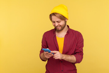 Wall Mural - happy trendy hipster guy in beanie hat and checkered shirt using cell phone, texting or reading mess