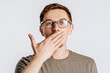 © Daria Lukoiko - Young handsome man with glasses a brunette beard posing on isolated gray background. Place for advertising. The surprised man is shocked and covers his mouth with his hand. Bad surprise news.