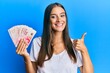 © Krakenimages.com - Young hispanic woman holding israel shekels smiling happy and positive, thumb up doing excellent and approval sign