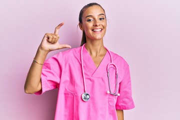 Wall Mural - Young hispanic woman wearing doctor uniform and stethoscope smiling and confident gesturing with hand doing small size sign with fingers looking and the camera. measure concept.