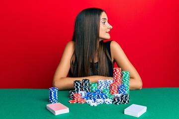 Wall Mural - Young hispanic woman sitting on the table playing poker looking to side, relax profile pose with natural face with confident smile.