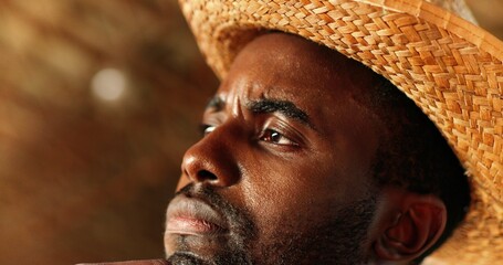 Portrait of African American young handsome man in hat thinking with serious face and looking at side in shed. Close up of face of male farmer considering something. Indoor.