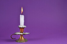 Very Old Rustic Copper Candlestick With White Paraffin Burning Candle And Copy Space On Purple Background