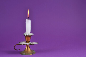 Very old rustic copper candlestick with white paraffin burning candle and copy space on purple background