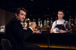 © Anna Kosolapova - a handsome man poses at the bar with a cocktail in his hands against the background of the bartender and bottles.