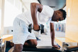 © twinsterphoto - Side view of African American man in sportswear leaning on bench and doing row exercise with dumbbell during training in gym.