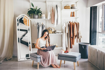 Smiling young lady with modern laptop talks on mobile phone in comfortable room at home.