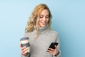 Young blonde woman wearing a sweater isolated on blue background holding coffee to take away and a mobile