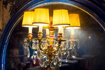 a candelabrum with a yellow lampshade with glass elements on an old mirror with a worn blue wooden frame, a wall lamp in a dark retro interior shines with warm light closeup side view, nobody.