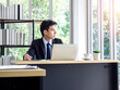 © tete_escape - Young Asian businessman in suit working with laptop computer in office with green tree background outside.