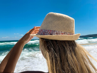 Young blond woman with hat standing on the beach at summer day watching in the distant emerald sea and blue sky in background