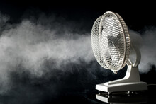 Side View Of A White Electric Desktop Fan On A Black Background And Reflective Floor With Visible Fog Or Mist Blowing Through. Concept Of Freshness With A Fan