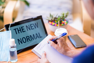 Woman writes by hand from home on a notepad - surgical mask due to covid-19 and tablet with message for the new normal concept on the wooden table