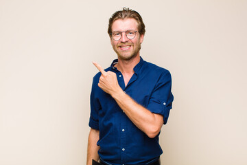 Wall Mural - blonde adult caucasic man smiling cheerfully, feeling happy and pointing to the side and upwards, showing object in copy space