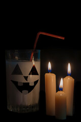 Wall Mural - In the dark, against a black background, there is a glass with an eerie face, with a white drink illuminated by the flame of three sevches. idea - design of drinks for a party. Halloween.