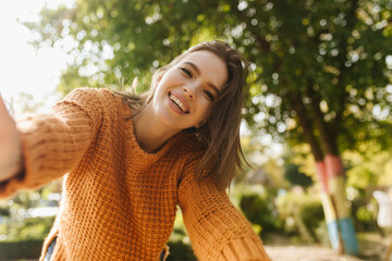 Wall Mural - Refined woman in trendy sweater having fun in park. Adorable girl taking selfie on blur nature background.
