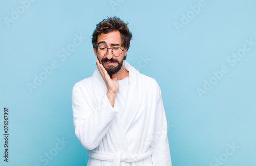 Fotomural young bearded man wearing a bath robe holding cheek and suffering painful tootha