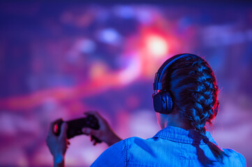 Wall Mural - The girl gamer is actively playing games with her back, against the background of a 4k TV, or a larger screen. Gaming, streaming, joystick and passing the game