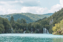 Plitvice Lakes Breathtaking Waterfalls View. Wild Blue Green Heart Of Mountains Sheltered By Rocky Walls Of Mountains. Mountain Waters Gathered In White Foamy Waterfalls Overflows In Beautiful Lake