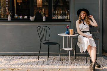 Poster - A cute, elegant girl sits on a chair in a street cafe and touches her hat