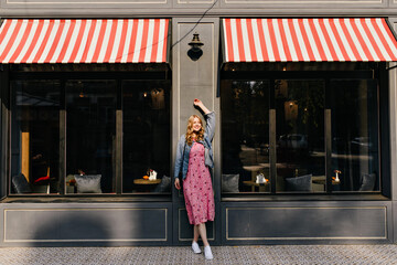 Wall Mural - horizontal photo in full growth of a happy blonde with her right hand raised in a denim jacket and white sneakers against a street cafe