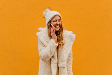 Wall Mural - Young joyful girl talking on phone and closes her eyes with laughter on orange background