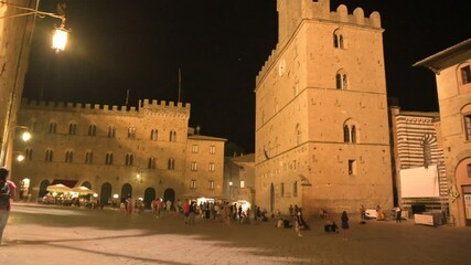 Wall Mural - Volterra,Tuscany,Italy.August 2020. Night footage in the magical atmosphere of Piazza dei Priori. Three-quarter view of the homonymous building, the wind moves the flags slightly, people in the square