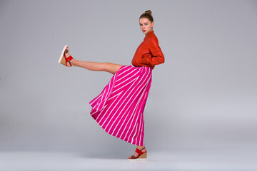 Young woman posing in full growth. Clothes - a long red skirt with a wide white stripe, and orange bomber jacket, wedge sandals. She makes a high leg swing.